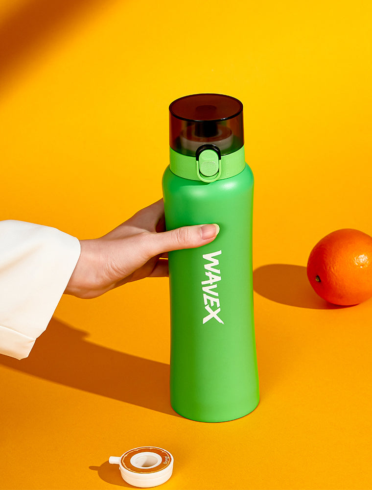 A hand grips a green 550ml steel water bottle next to a bright orange, highlighting the honey peach taste experience.