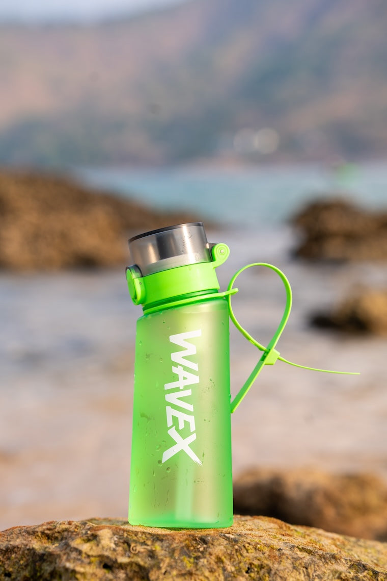 A green 550ml water bottle with a handle sits on the beach, featuring a refreshing honey peach flavor